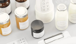  A collection of cosmetic or skincare products, curated by a Kreativagentur, is displayed on a white surface. The items feature jars and bottles with minimalist white labels, printed with words like Apfel, Gen Mai, and Vitalbad. Nearby lies a sleek metal tool or accessory.